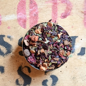 May include: A close-up of a bowl filled with a blend of dried fruit, herbs, and spices. The mixture includes red berries, cinnamon sticks, apple pieces, and green leaves.