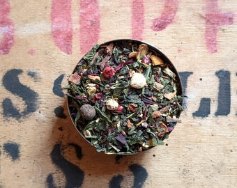 May include: A close-up of a metal bowl filled with a blend of green, red, and white tea leaves. The tea blend includes dried fruit, flowers, and spices.