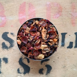 May include: A close-up of a metal bowl filled with a blend of dried fruit, spices, and herbs. The mixture includes red and orange fruit pieces, brown cinnamon sticks, and other brown and beige ingredients.
