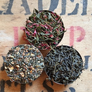 May include: Three small bowls filled with different types of loose leaf tea. The bowls are arranged in a triangle shape on a wooden surface. The tea in the top bowl is a green tea with pink flower petals. The tea in the left bowl is a blend of green tea with spices and dried fruit. The tea in the right bowl is a dark green tea.