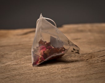 May include: A single-serve tea bag filled with dried rose petals and other herbs. The tea bag is made of a clear, mesh material and is resting on a wooden surface.