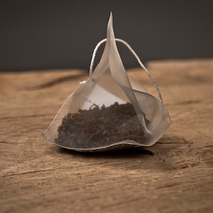 May include: A single-serve tea bag filled with loose leaf tea. The tea bag is made of a white mesh material and is shaped like a pyramid. The tea bag is sitting on a wooden surface.