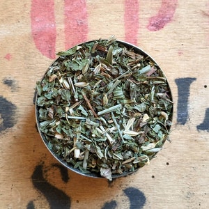 Könnte beinhalten: Nahaufnahme einer kleinen Metallschale gefüllt mit einer Mischung aus getrockneten Kräutern, darunter grüne Blätter, Stängel und kleine Blütenknospen.
