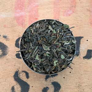 May include: A close-up of a small metal bowl filled with loose leaf green tea. The tea leaves are a mix of dark green and light green, with some white tips.