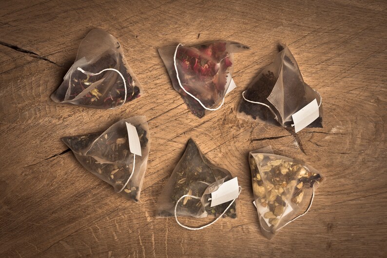 May include: Six triangular tea bags filled with different types of tea leaves. The tea bags are arranged in a circle on a wooden surface.
