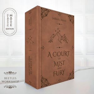 Könnte beinhalten: Ein braunes Buch mit dem Titel "A Court of Mist and Fury" von Sarah J. Maas. Das Buch hat dekorative Eckendesigns und Bergillustrationen. Das Cover enthält auch den Text "Matte Classic Edition" und das Logo "Beetles Workshop".