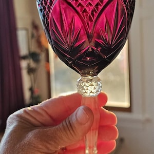 May include: A ruby red crystal wine glass with intricate cut glass patterns. The bowl features a diamond and fan design, with a clear, faceted stem and base. The glass is held in a hand, showcasing its elegant form.