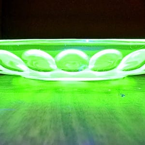 May include: A glowing green glass dish with a scalloped edge and a series of oval-shaped indentations. The dish is illuminated, creating a bright, neon-like effect. The dish is likely vintage or antique.