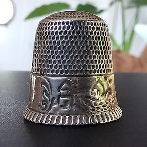 May include: A silver-colored metal thimble featuring a textured honeycomb pattern on the upper dome and a smooth band around the base. The base band is decorated with embossed relief designs including a small house and a sailboat motif.
