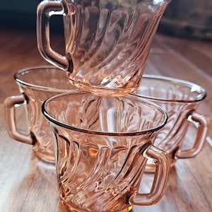 May include: Four vintage pink glass teacups with handles. The cups have a swirled design and are stacked on a wooden surface. The cups are a light pink color and appear to be made of glass.