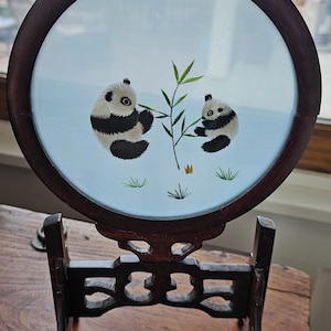 May include: A round, decorative wooden display featuring an embroidered panda bear scene. Two pandas are depicted eating bamboo on a light blue background. The dark brown frame and stand add a touch of elegance to this unique piece.