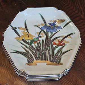 May include: An octagonal ceramic plate with a cream-colored base featuring a painted design of blue, yellow, and orange iris flowers, green leaves, and a small bird. The plate has a gold-trimmed edge and subtle gold leaf accents throughout.