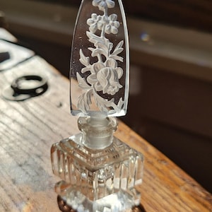May include: A clear glass perfume bottle with a square, vertically ribbed base and a pointed, triangular stopper featuring an intricate, frosted floral relief carving. The glass is transparent, catching the light to highlight the detailed petals.
