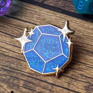 May include: A blue and gold enamel pin shaped like a faceted gem, with gold star accents. The gem has a glittery, iridescent finish. Two multi-sided dice are in the background, one purple and one teal, with white numbers. The pin is on a wooden surface.