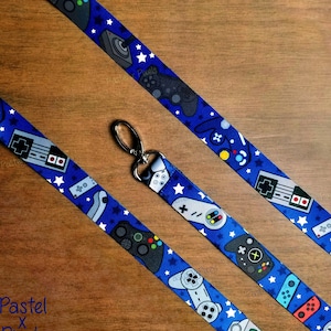 May include: Blue lanyard with a pattern of white stars and various video game controllers. The lanyard has a silver-colored clasp and the text "Pastel x Pixels" in the lower left corner.