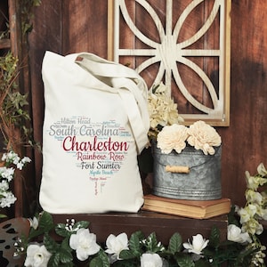 May include: A white canvas tote bag with a word cloud design featuring the text "Charleston, South Carolina" and other related locations. The bag is sitting on a wooden shelf with flowers in a metal bucket.