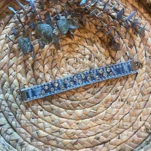 Puede incluir: Una pulsera de mezclilla con pedrería plateada. La pulsera rectangular presenta una fila central de pedrería. Descansa sobre una bandeja circular, tejida, de color beige, adornada con hojas de eucalipto secas. La pulsera mide unos 15 cm de largo.