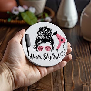 May include: A white, round coaster featuring a black and pink graphic design. The design includes a silhouette of a woman's updo hairstyle, pink leopard print glasses, a comb, and a hairdryer. The words "Hair Stylist" are written below.