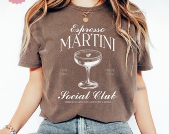 May include: A brown t-shirt with a white graphic design that says "Espresso Martini" with a martini glass illustration and the text "Tini Time" and "Since 1974". The graphic also includes the text "Social Club" with the tagline "Stirred, Shaken, and Absolutely Awake".