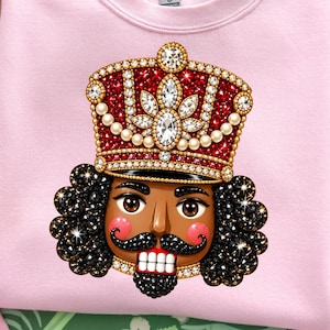 May include: A light pink crewneck sweatshirt featuring a graphic of a nutcracker head wearing a red and gold jeweled crown. The nutcracker has dark skin, black curly hair, a black mustache, and a white-toothed grin with red cheeks.