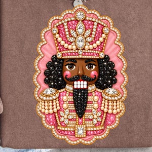 May include: A brown crewneck sweatshirt featuring a graphic of a nutcracker decorated with pink and gold rhinestones. The nutcracker has a black mustache, a tall hat, and a uniform with epaulets. A silver chain necklace rests on the collar.