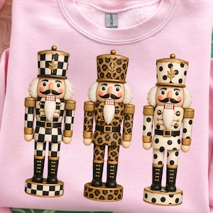 May include: A light pink crewneck sweatshirt featuring a graphic print of three nutcracker figures. The first nutcracker has a black and white checkered pattern, the second has a brown leopard print, and the third has a black polka dot pattern.