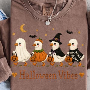 Puede incluir: Una sudadera marrón de cuello redondo con un gráfico de cuatro patos con disfraces de Halloween, cada uno sosteniendo un cubo de calabaza. Los patos están vestidos de fantasma, calabaza, bruja y esqueleto. El texto "Halloween Vibes" aparece debajo.