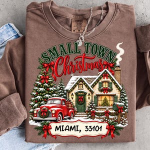 Op de afbeelding: Een bruin sweatshirt met lange mouwen en een afbeelding van een rode vrachtwagen, een versierd huis en een kerstboom. De tekst luidt Small Town Christmas en Miami, 33101. Het ontwerp bevat groene dennenbomen, rode strikken en sneeuw op een bruine stof.