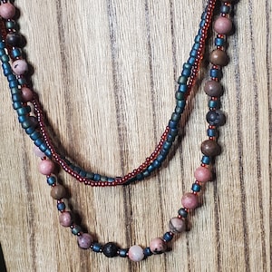 May include: A layered necklace with brown, pink, and teal beads. The necklace is made of small seed beads and larger round beads.