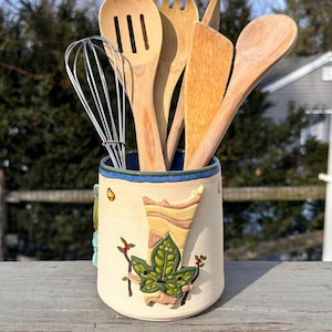 Puede incluir: Un recipiente de cerámica para utensilios con base color crema y borde azul, decorado con un diseño de hojas y bayas verdes. Contiene una variedad de utensilios de cocina de madera, incluyendo un batidor, cucharas y una espátula.