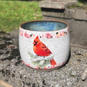 May include: A ceramic mug with a speckled white glaze featuring a vibrant red cardinal bird perched on a green pine branch. The rim is finished in a deep blue, and the top and bottom edges are decorated with a soft pink and red watercolor-style band.