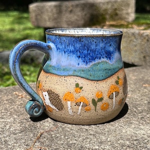 May include: A ceramic mug with a blue and turquoise glazed rim and handle. The mug features a tan base with hand-painted mushrooms, berries, and a hedgehog design. The mug is suitable for hot or cold beverages.