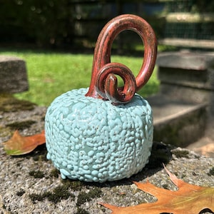 May include: A ceramic pumpkin sculpture featuring a textured, light blue, bubbly glaze on the round body and a smooth, glossy, dark reddish-brown curved stem that loops into a spiral at the top, creating a distinct contrast in color and finish.