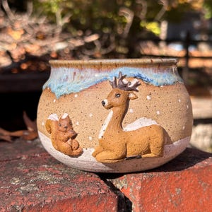 May include: A ceramic planter with a tan base, featuring a deer and squirrel relief design. The rim has a blue and brown glaze, with a snowy landscape effect. The planter is sitting on a red brick surface.