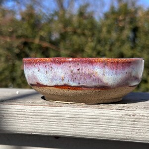 May include: A small, handmade ceramic bowl with a speckled, glazed interior. The rim is a deep red, transitioning to a light blue and purple center. The base is unglazed, showing the natural tan clay. A decorative piece for the home.