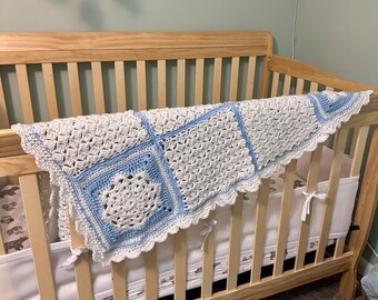 May include: A handmade, white and light blue crochet blanket draped over a wooden crib. The blanket features a square pattern with a floral design in the center of each square. The edges are scalloped, and the blanket is made of soft yarn.