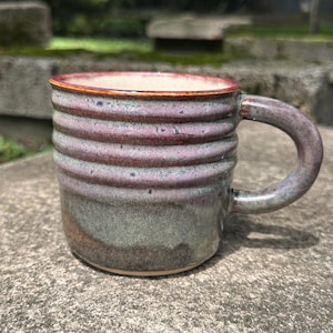 Könnte beinhalten: Ein Keramikbecher mit gerippter Textur in der oberen Hälfte und einer gesprenkelten violetten und rosafarbenen Glasur. Der untere Teil geht in ein glattes, erdiges Grün und Braun über. Er verfügt über einen stabilen, geschwungenen Henkel und einen rotbraunen Rand.