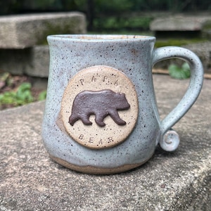 May include: A light blue speckled ceramic mug with a rounded handle and a circular medallion on the front. The medallion features a dark brown embossed bear silhouette in the center with the text PAPA BEAR stamped around the bear graphic.