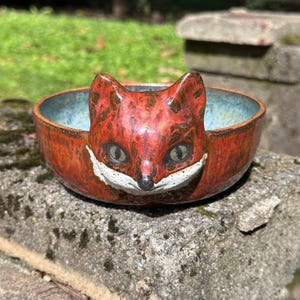 May include: A round ceramic bowl featuring a sculpted fox head on the front rim. The fox has a reddish-brown coat with dark brown patches, a white muzzle, and green eyes. The interior of the bowl is finished in a mottled light blue glaze.