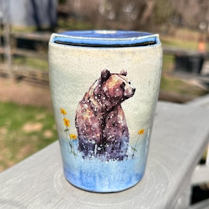 May include: A ceramic tumbler with a watercolor painting of a brown bear. The bear is surrounded by yellow flowers and blue water. The top and bottom of the tumbler are blue.