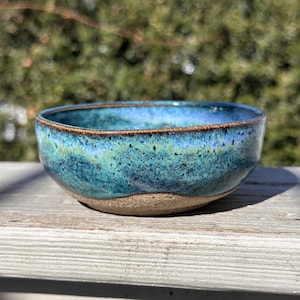 May include: A handmade ceramic bowl with a speckled turquoise glaze. The bowl has a rounded shape with a brown rim and a natural, unglazed base. The interior has a gradient of blues and greens.