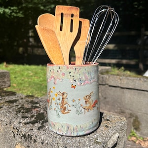 May include: A cylindrical ceramic utensil holder with a light green glaze and a decorative print of brown rabbits, yellow flowers, and a small orange butterfly. The holder contains three wooden kitchen utensils and a metal wire whisk.