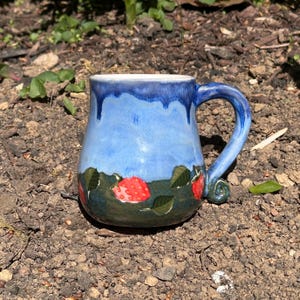 Puede incluir: Una taza de cerámica azul con un glaseado de goteo y un asa. La taza está decorada con un paisaje verde y marrón con fresas rojas.