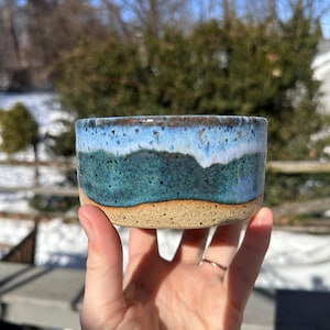 May include: A handmade ceramic bowl with a textured, layered design. The bowl features a gradient of colors, including light blue, teal, and a tan base. The bowl is held in a hand, showcasing its size and shape.