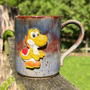 Peut inclure: Mug en céramique fait main avec un personnage Yoshi jaune en relief. Le mug présente un glaçage moucheté bleu et marron. La poignée est incurvée. Un objet de collection idéal pour les amateurs de jeux vidéo.