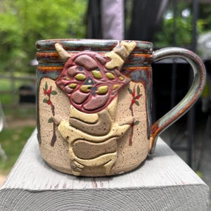 May include: A ceramic mug with a speckled beige base and a dark brown, green, and blue glazed rim. The front features a raised, sculpted relief of a stylized creature with a brown head and tan body, flanked by two painted branches with red berries.