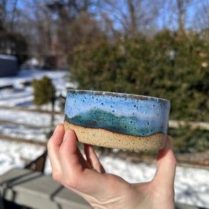 May include: A handmade ceramic bowl with a blue and green mountain design. The bowl has a tan base and a speckled blue top. The bowl is held in a hand, with a blurred outdoor background.