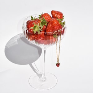Könnte beinhalten: Eine klare Champagnerflöte aus Glas, gefüllt mit frischen roten Erdbeeren. Eine goldene Kettenkette mit einem Erdbeeranhänger hängt am Rand des Glases.