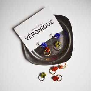 Könnte beinhalten: Ein Set handgemachter Ohrringe mit kleinen, blütenförmigen Anhängern aus bunten Perlen. Die Ohrringe werden auf einem silbernen Tablett mit einer weißen Karte präsentiert, auf der "HANDMADE JEWELRY VÉRONIQUE" steht.
