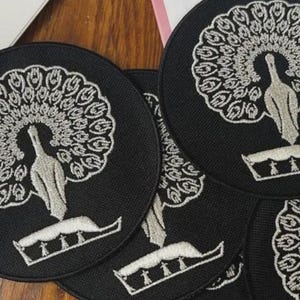 Pu&ograve; includere: Una collezione di toppe circolari in tessuto nero con un ricamo raffigurante un pavone color argento. Ogni toppa presenta un pavone stilizzato con le piume della coda spiegate a ventaglio circolare sopra un elemento di base decorativo.